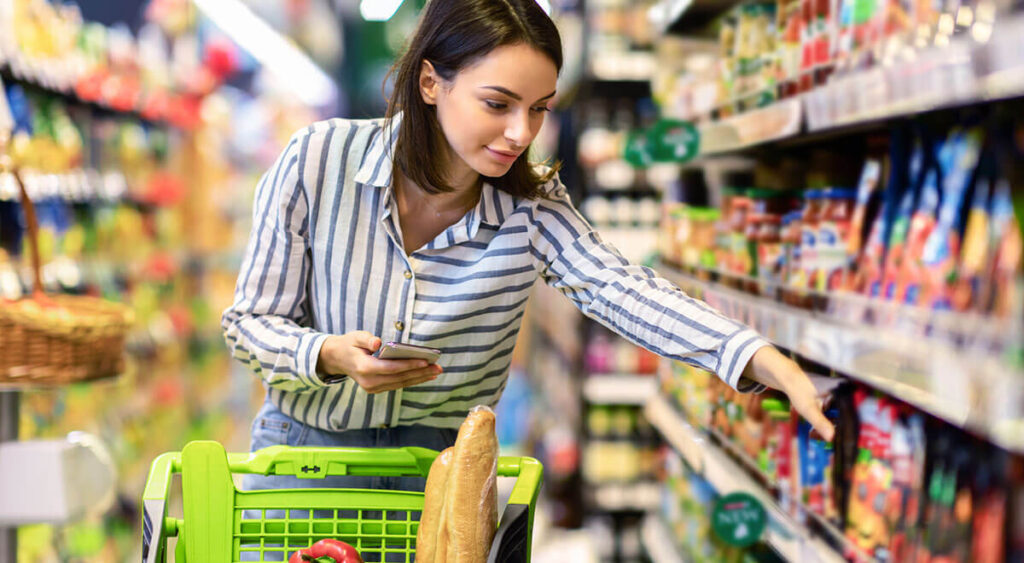 A woman gazes thoughtfully at a cart overflowing with groceries in a bright, bustling store.
